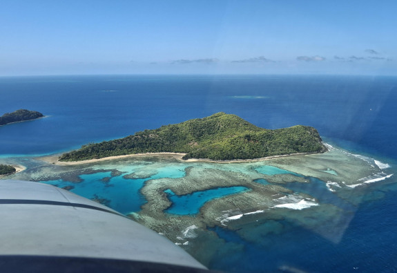 Image of an island taken in the sky, you can see part of the aircraft to the left of the photo. The island looks tropical with very blue water. There is blue sky. 