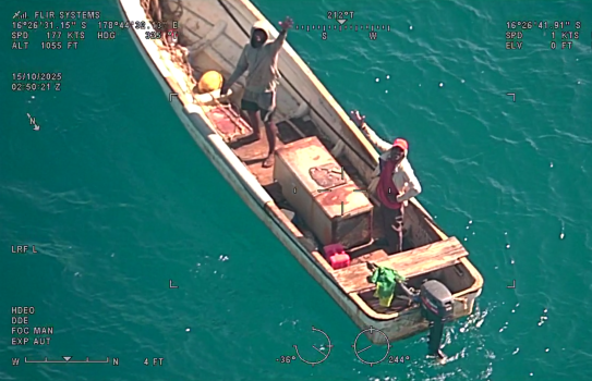 Photographed from an aircraft above, two men look up and wave from a small boat at sea.