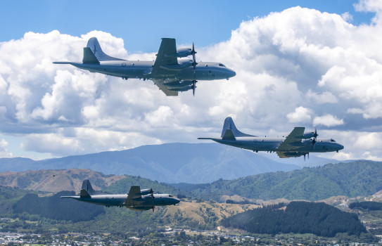 Three grey Orion aircraft fly in a 'V' formation over suburbia, with rolling hills, clouds and a slither of blue sky in the background.