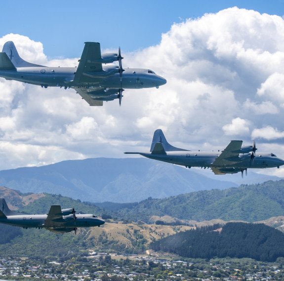 Three grey Orion aircraft fly in a 'V' formation over suburbia, with rolling hills, clouds and a slither of blue sky in the background.