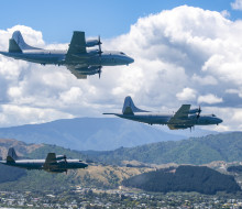 Three grey Orion aircraft fly in a 'V' formation over suburbia, with rolling hills, clouds and a slither of blue sky in the background.