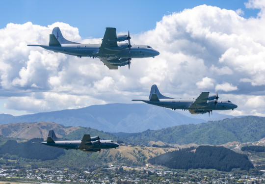 Three grey Orion aircraft fly in a 'V' formation over suburbia, with rolling hills, clouds and a slither of blue sky in the background.