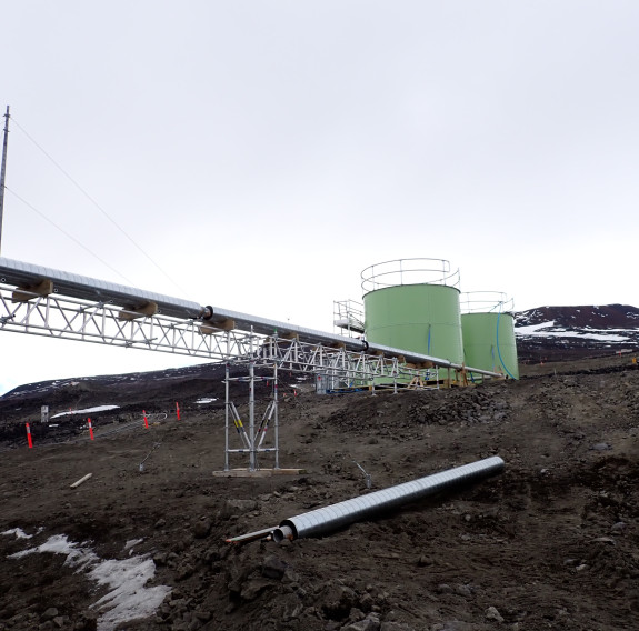 Two large green water containers sit on the side of a rocky hill with the peak in the background covered in snow. There are insulated pipes that go from the water containers on top of scaffolding.