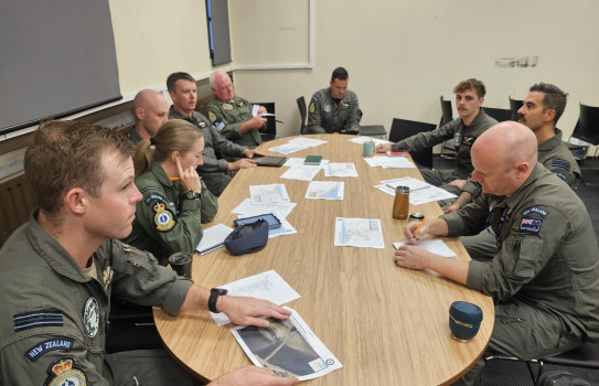 The crew of a Poseidon aircraft have a discussion around a wooden oval table. Personnel are in uniform and paperwork and various beverages are strewed across the table.