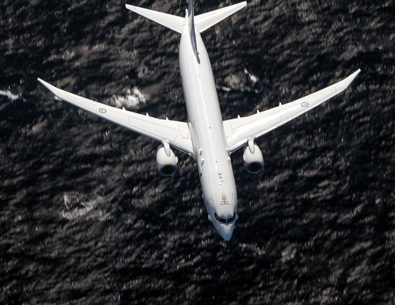 Large grey aircraft with a single jet on each wing flies over the ocean.