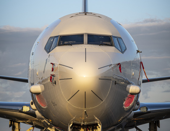 A nose-on perspective of the P-8A Poseidon. The large grey aircraft glows yellow as the sun reflects off the nose. In the background, low cloud meets blue skies.