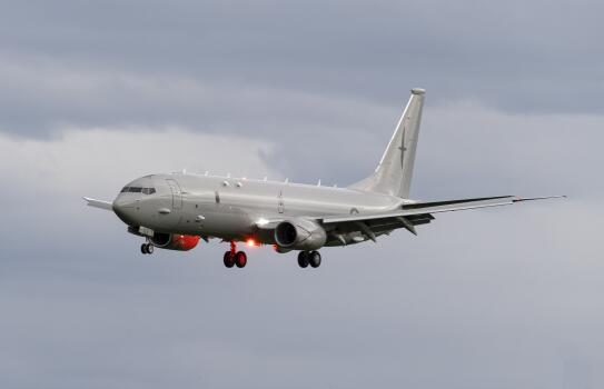 A RNZAF P-8A Poseidon in flight in front of clouds. A red beacon lights the underneath of the aircraft as the landing gear is deployed.