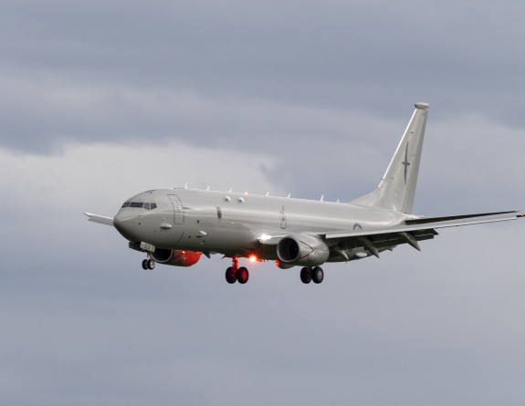 A RNZAF P-8A Poseidon in flight in front of clouds. A red beacon lights the underneath of the aircraft as the landing gear is deployed.