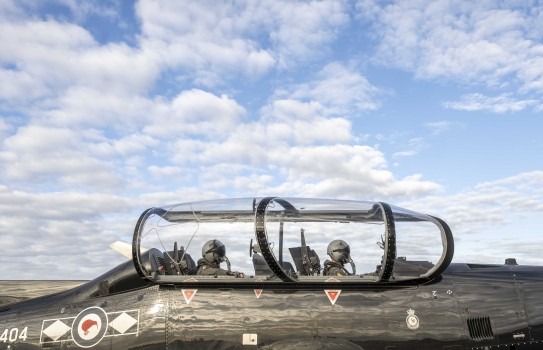 Two pilots can be seen sitting in a black small aircraft, one behind the other wearing helmets that are connected to oxygen.  The cockpit is lifted up on the tarmac.