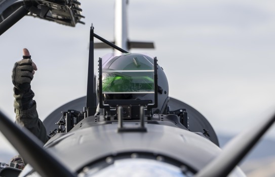 A pilot sits in an aircraft and looks to his right whilst giving a thumbs up, his face obscured by a screen in front of him
