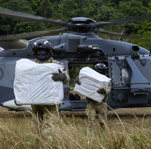 Two RNZAF aviators wearing military uniform and flight helmets, carry large white wrapped packages off a NH90 helicopter to a remote community.