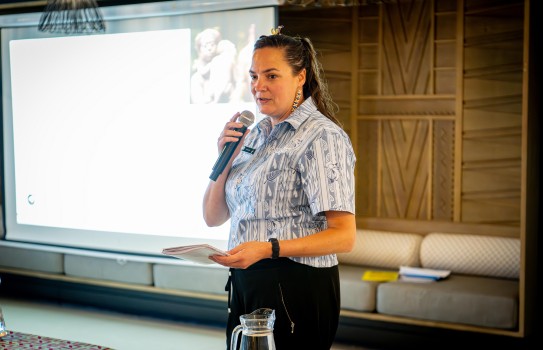A woman stands in front of a project and talks into a microphone while holding a booklet.