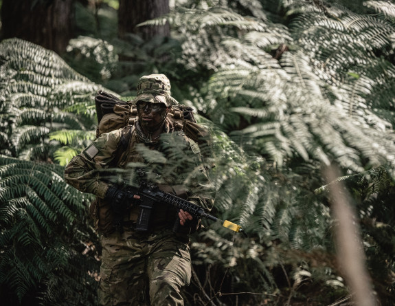 A soldier crouches in the bush, carrying a weapon. The soldier is quite camouflaged. 