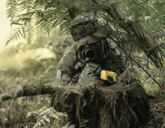 A soldier crouches in the bush, carrying a weapon. The soldier is quite camouflaged. 