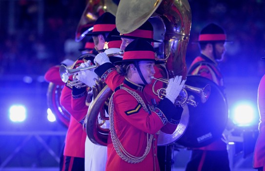 Army band members wearing red and black uniform play a variety of brass instruments in front of a line of blue lights.