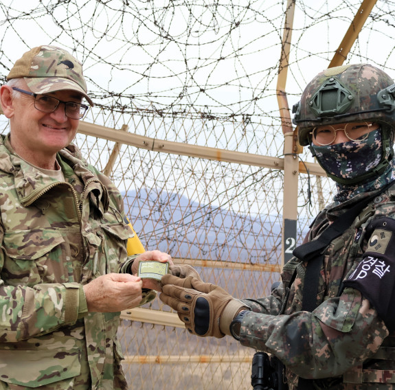Two soliders was a New Zealand patch in front of a fence with barbed wire on top.