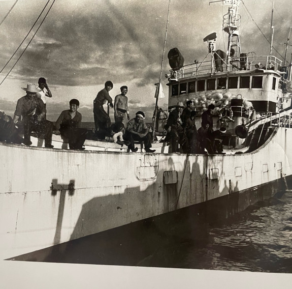 A sepia historic photo of a Japanese fishing vessel with fishermen sitting on the edge of the vessel.
