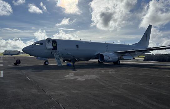 RNZAF P-8A Poseidon at Guam on the flight line. There is some blue sky as well as clouds. The front aircraft door is open and the stairs down. There is someone in a high vis in doorway and someone walking around the aircraft. 