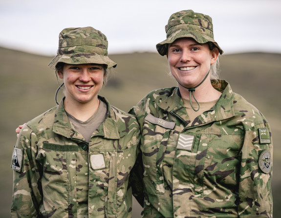 Two solders in uniform stand side-by-side, smiling at the camera. The background is blurred. 