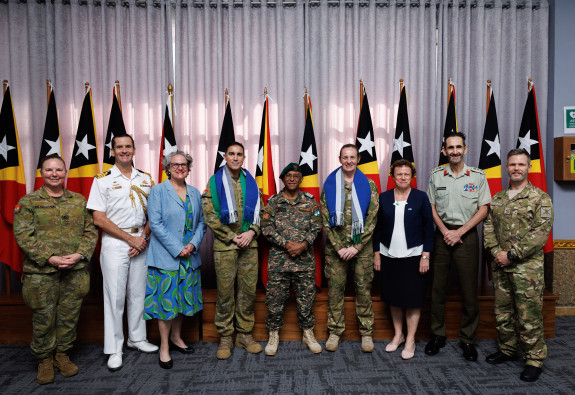 Nine people stand in a row - all in military uniform except two. They stand with their hands in front of them and they are all standing in front of a row of Timor-Leste flags. 