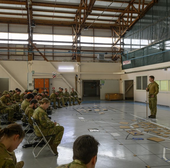 A soldier stands on the right in front of soldiers that are seated in a half circle in a gymnasium,