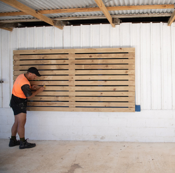 A man wearing a high visibility t-shirt, black shorts, hat and boots installs wooden protective slats on a window of a community hall.