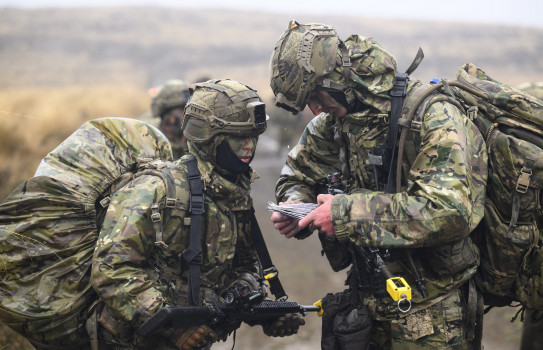 Two soldiers in full wet weather Army uniform and large packs look at documentation in the rain on exercise.