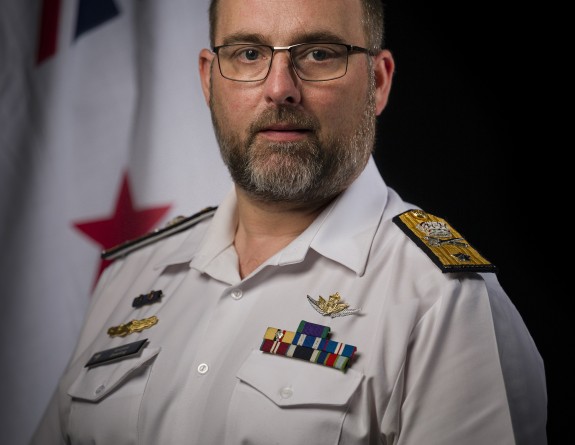 A man in Navy uniform, a white short-arm shirt with rank slides looks at the camera. He has a short beard and is wearing glasses. In the background hangs the RNZN ensign.