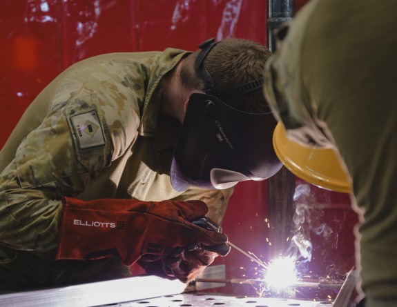 Two soldiers weld a steel grate, both wear protective safety equipment of orange gloves and welding masks to protect themselves from the welding arc 