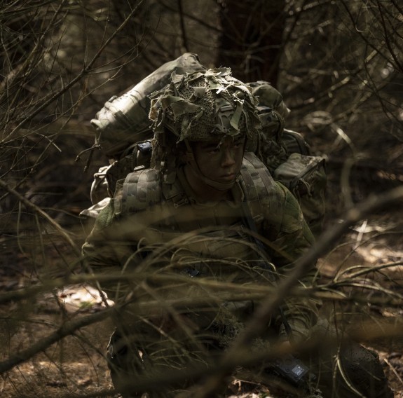 A soldier sits waiting amongst the forest canopy, camouflaged maintaining a visual of their surrounds.