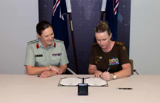 Two personnel in New Zealand and Australian military uniforms sign a document in front of their corresponding countries flags.