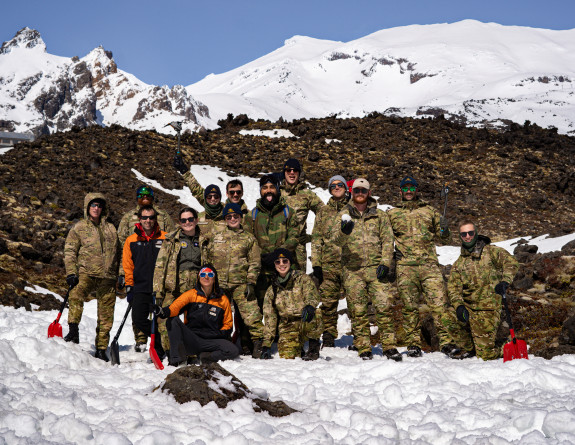 A group photo of personnel standing on the mountain looking at the camera. Snow is all around them as well as mountain rock. There are plenty of happy faces. 