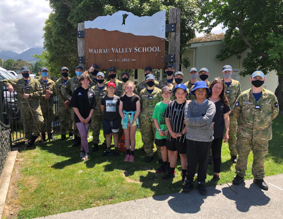 The officer cadets with some of the students and staff from Wairau Valley School. The uniformed personnel have masks on and it is a sunny day. 