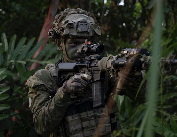 A soldier looks down the barrel of their weapon, dressed in uniform and also in the jungle.
