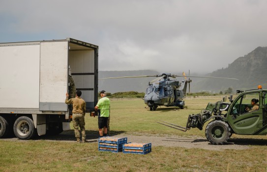 Three people are unloading bread of the back of a truck whilst a military forklift vehicle awaits.  Bread pallets are stacked in the foreground whilst a stationary grey military helicopter is in the background.