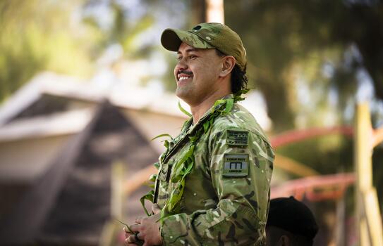 Person in camouflage uniform and cap standing outdoors with a leafy garland around the neck, with tents and trees blurred in the background.