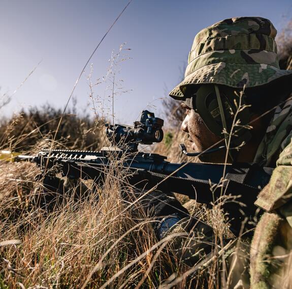 A soldier laying in the grass points their rifle away from the camera at an angle. The sun is low in the sky.