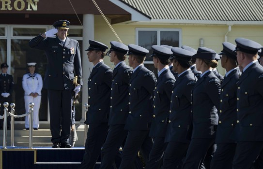 A senior officer stands on a podium saluting a body of Air Force personnel as they march whilst looking to their right. A naval person can be seen at the rear standing alongside an Air Force person looking on.