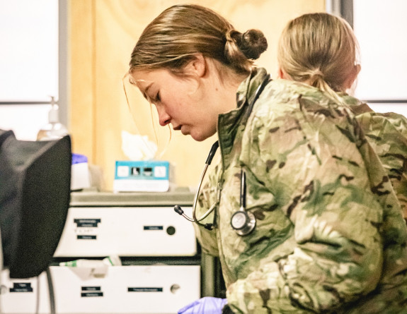 A solider in uniform assesses a patient during an Army medic exercise. In the background there is another soldier facing away. This soldier accessing the patient is side on to the camera. The patient is lying down. 