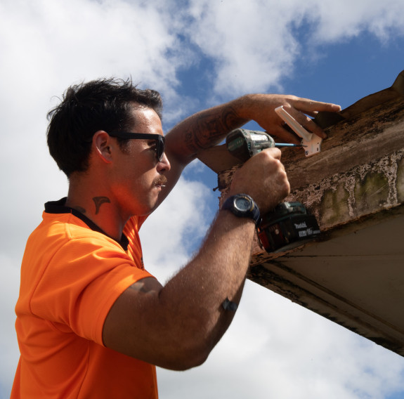 A man wearing a high visibility t-shirt and black sunglasses drills a bracket into where spouting will be installed on the exterior of a building.