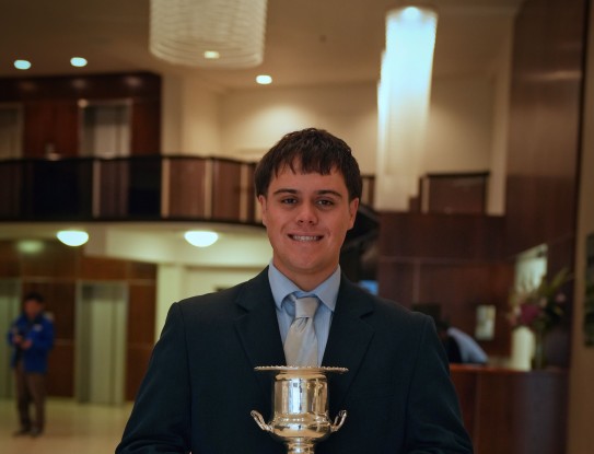 A man stands holding a trophy in a large auditorium, he wears a suit and stands proudly with a large smile on his face.