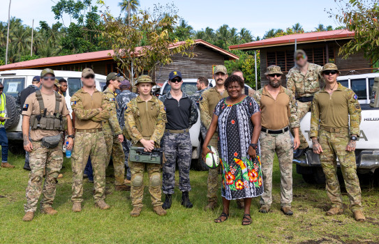 Soldiers stand with a local in front of white vehicles, buildings and tres