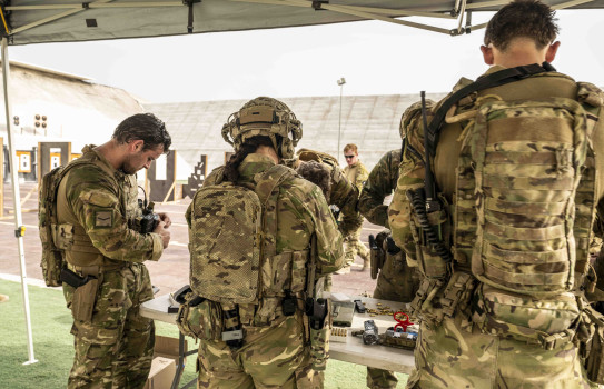 Three soldiers inspect weapons around a table, with a weapon range in the background.