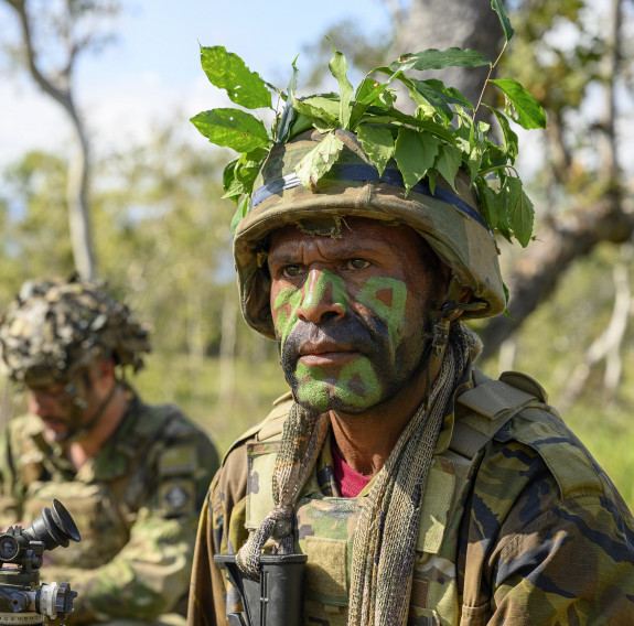 A PNGDF soldier wearing military uniform and camouflage paint sits next to a mortar with a NZ Army soldier in the background.