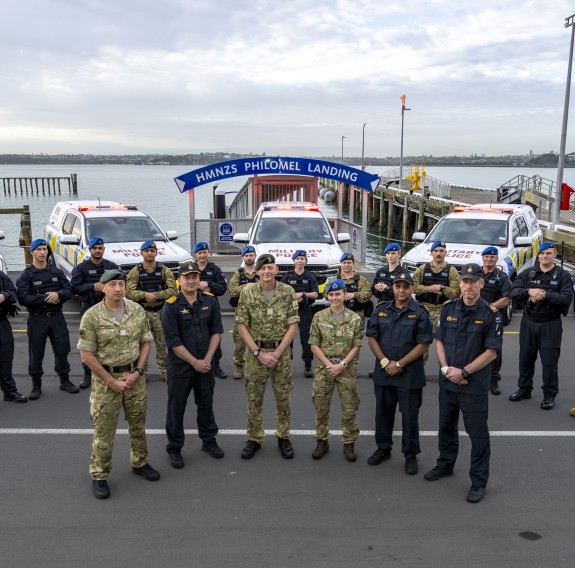 Soldiers, sailors and aviators stand in front of fleet of 4x4 vehicles by the edge of the water at Devonport Naval Base.