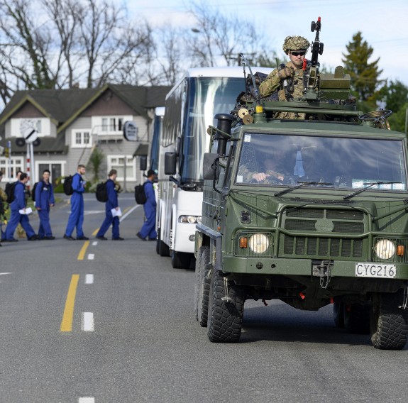 People in blue overalls board a coach as a green military vehicle is parked in front, with a soldier mounted on the gun on the roof.