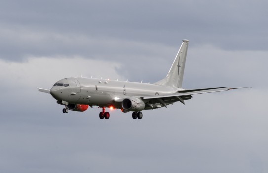 A grey aircraft with landing gear down in front of clouds.