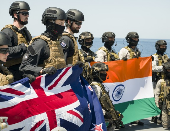 Military personnel in full combat attire each hold their countries flag while on a ship at sea.