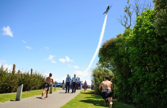 Senior RNZAF leaders walk in pairs down the path leading to the Turangawaewae. They are greeted by two toa who are flanking the path, each wielding taiaha and wearing customary piupiu. In the background an aeroplane steeply climbs skyward against the blue