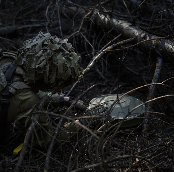 A soldier lays a metal circular object in the forest detritus, they are wearing webbing and camoflauge.